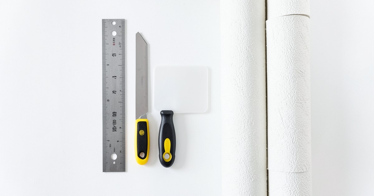 壁紙の張り替えに使用するカッター、定規、ヘラ、壁紙のロールが並んでいる様子